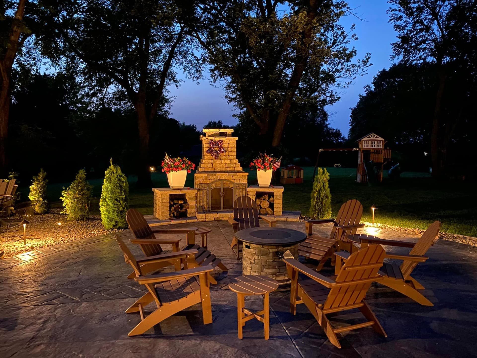 Stone patio with outdoor fireplace and Adirondack chairs illuminated by landscape lighting in Kansas City